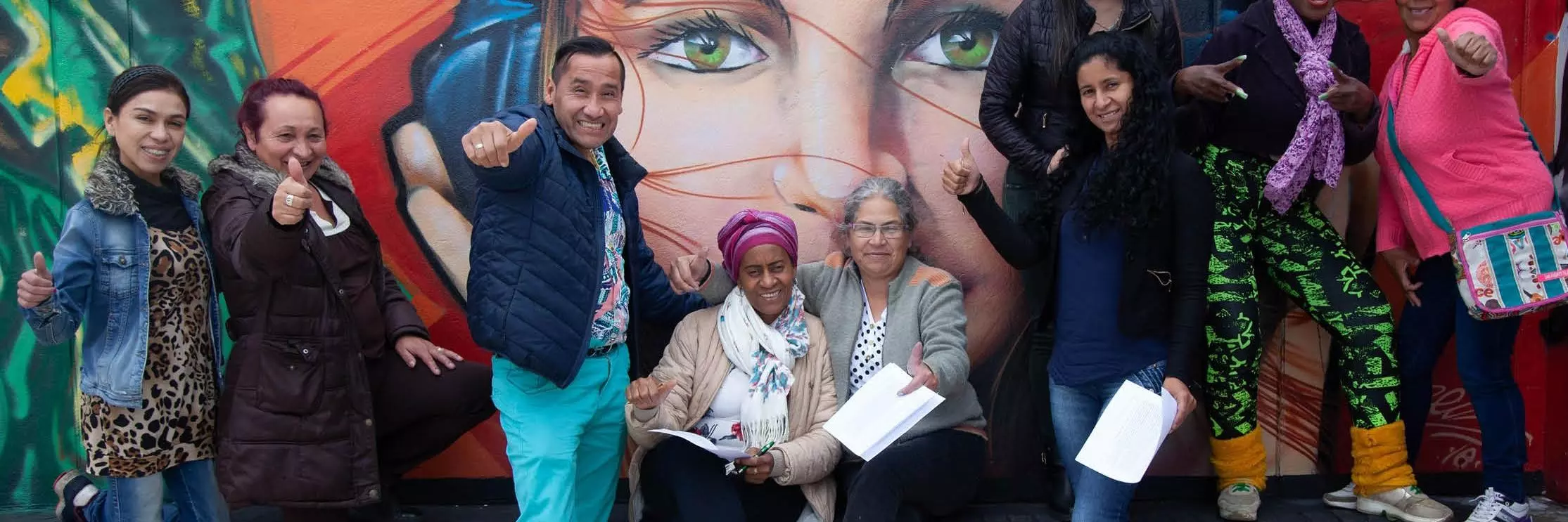 People hold their thumbs up to the camera in front of wall art in Bogota, Colombia